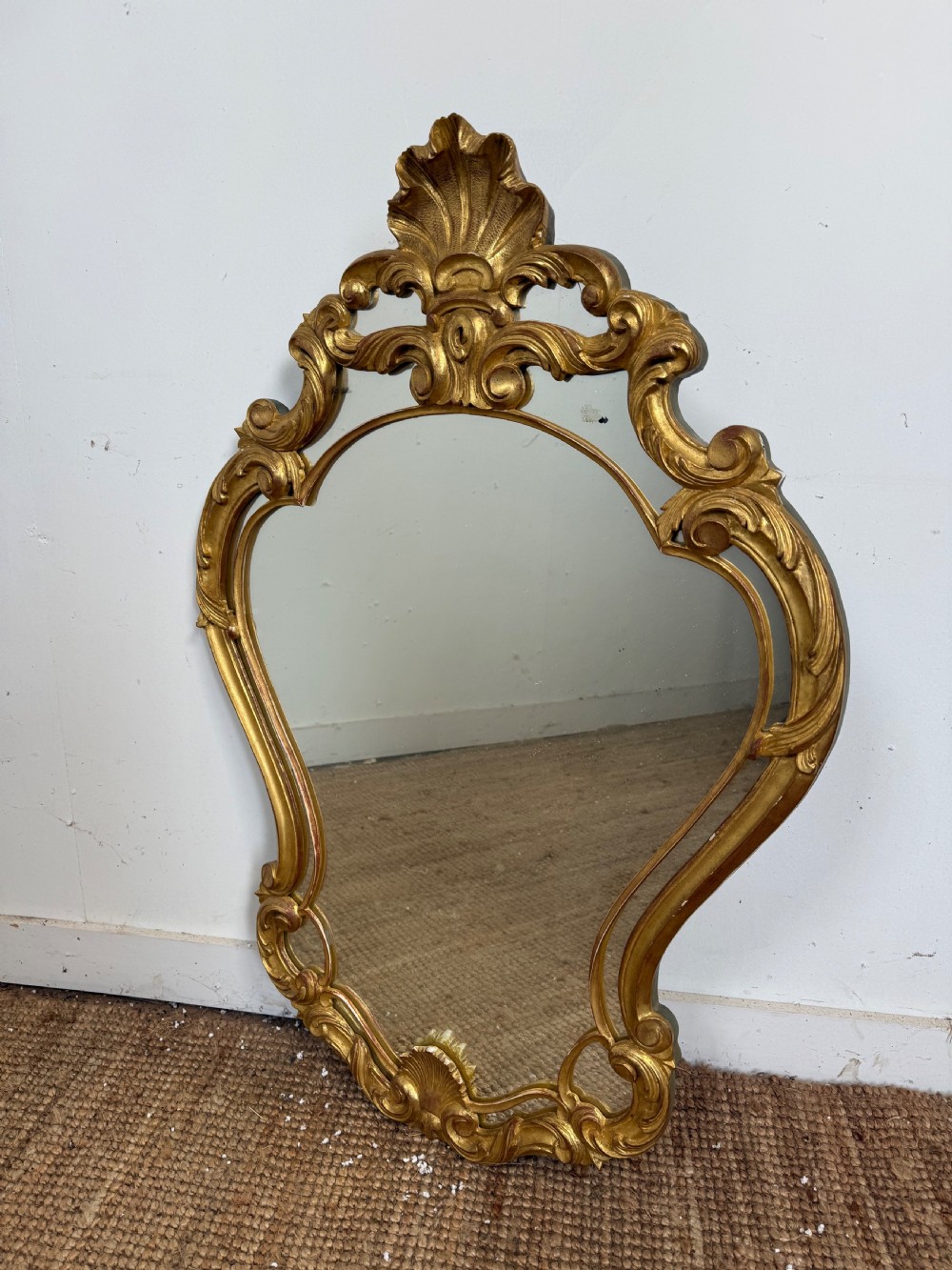 A French style carved giltwood wall mirror featuring an ornate shell crest and C-scroll border around an asymmetrical glass pane.