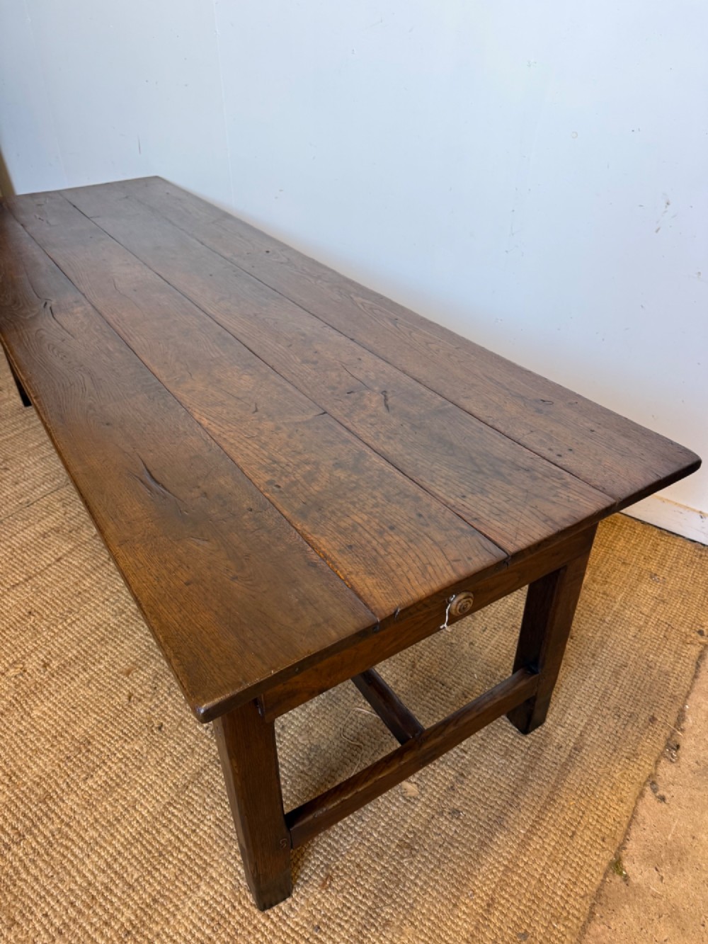 A long dark oak French farmhouse dining table with a plank top and an H-stretcher base structure. View X