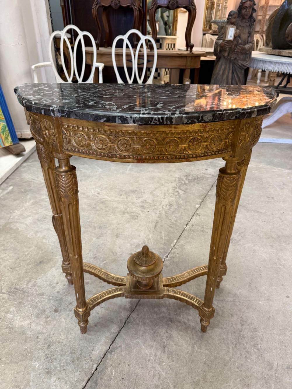 A late 19th-century French Louis XVI style demi-lune console table featuring a dark marble top, carved gilded legs, and a central urn finial.
