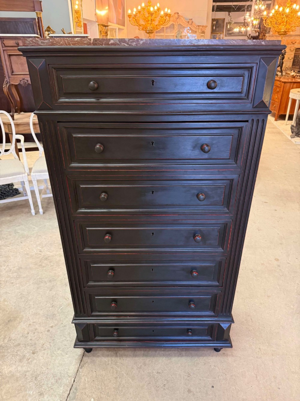 A tall narrow black ebonised chest of drawers featuring seven moulded drawers, turned wooden knobs, and a dark marble top.