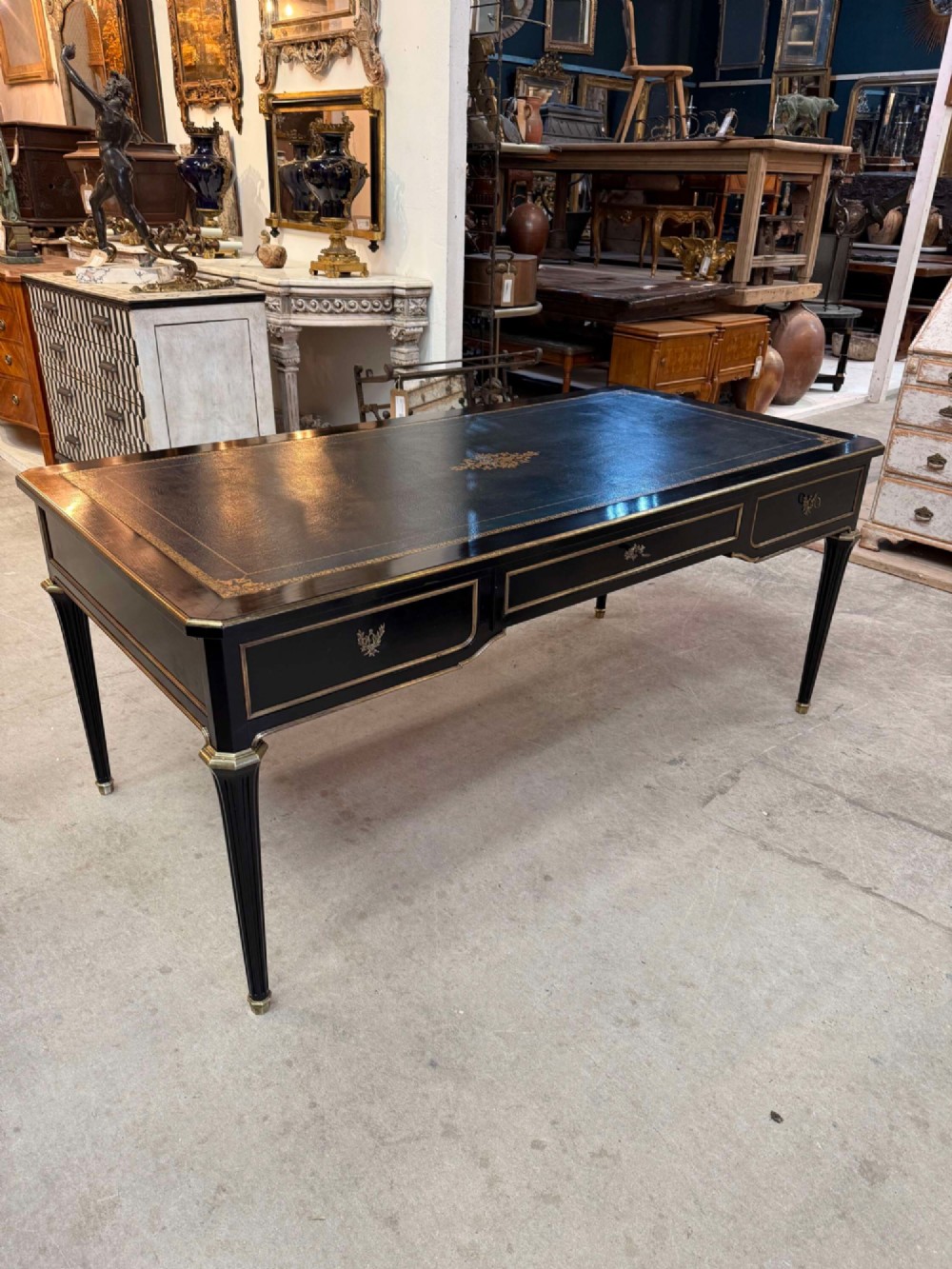 A 19th-century French ebonised writing desk with a black leather inset top, gilt-tooled border, three frieze drawers, brass mounts, and tapered fluted legs.