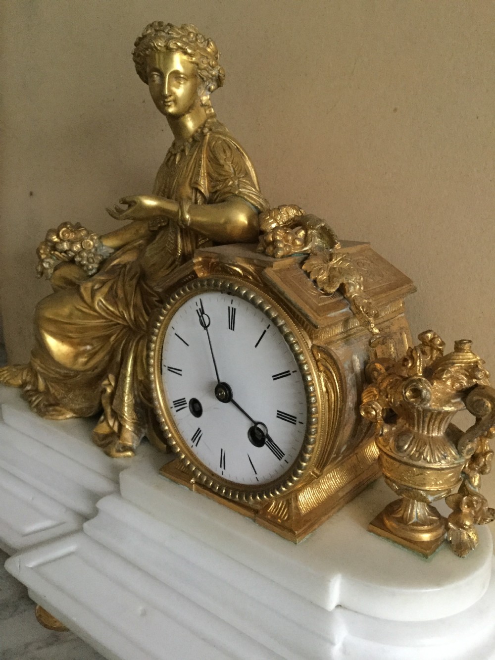 An antique French Empire mantel clock featuring a gilded bronze classical female figure leaning against a white enamel clock face set upon a stepped white marble base. View VI