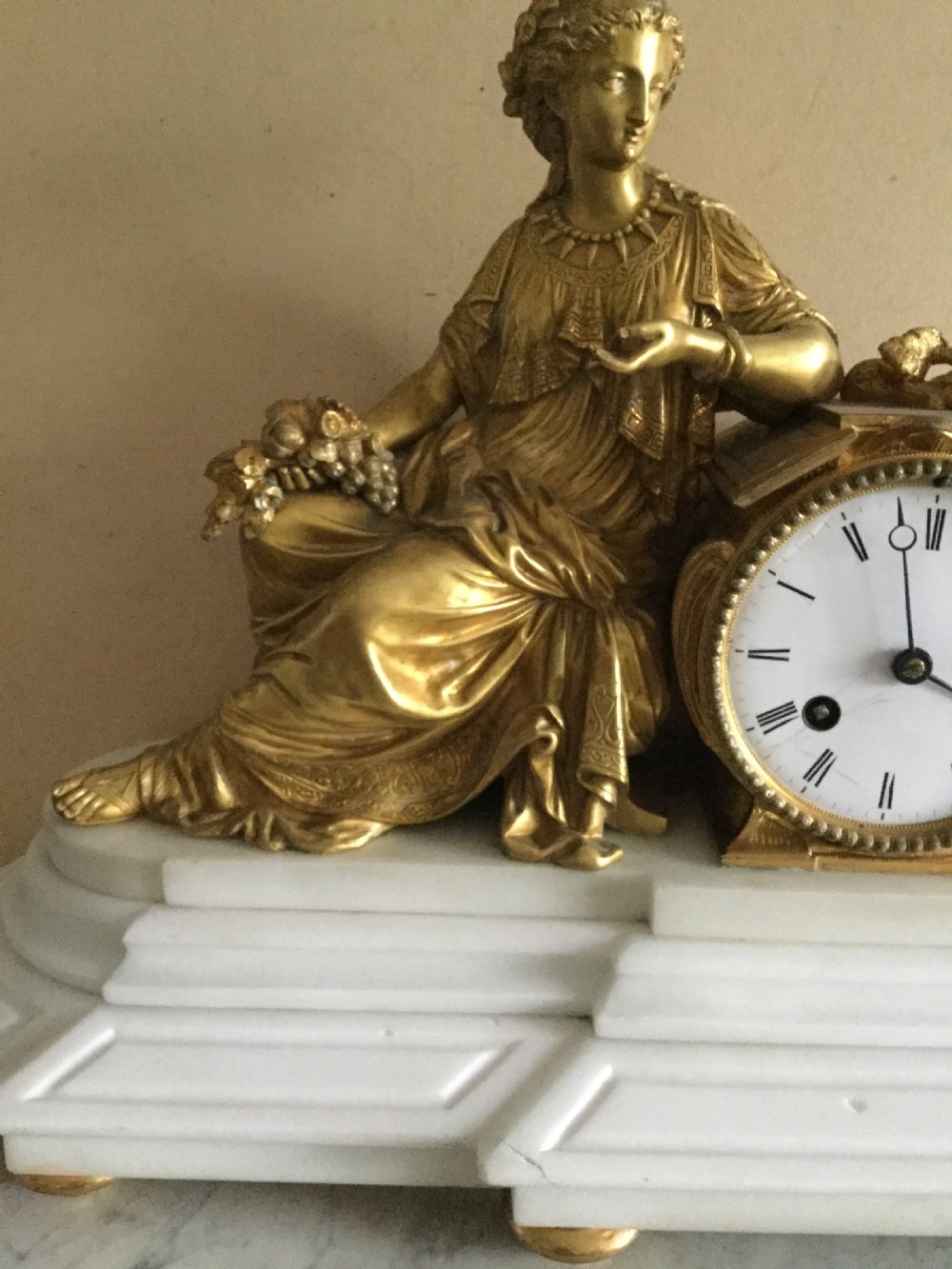 An antique French Empire mantel clock featuring a gilded bronze classical female figure leaning against a white enamel clock face set upon a stepped white marble base. View VII