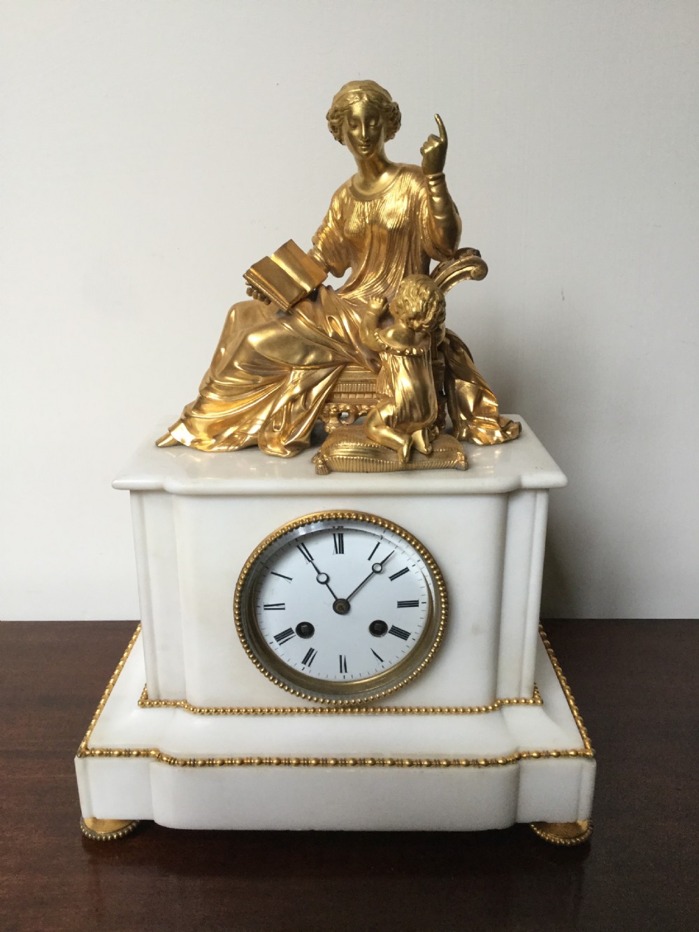 A French Neoclassical white marble mantel clock featuring a gilded bronze figural topper of a seated woman holding an open book alongside a cherub.