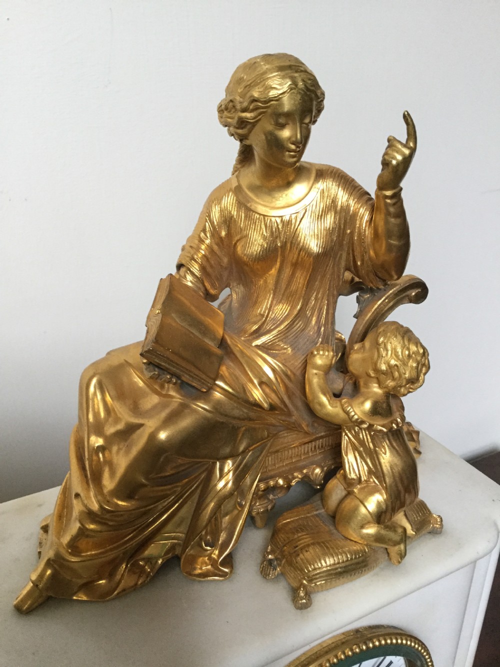 A French Neoclassical white marble mantel clock featuring a gilded bronze figural topper of a seated woman holding an open book alongside a cherub. View II