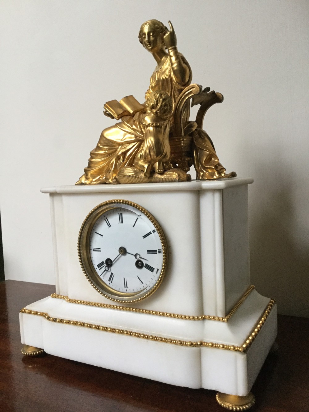 A French Neoclassical white marble mantel clock featuring a gilded bronze figural topper of a seated woman holding an open book alongside a cherub. View V