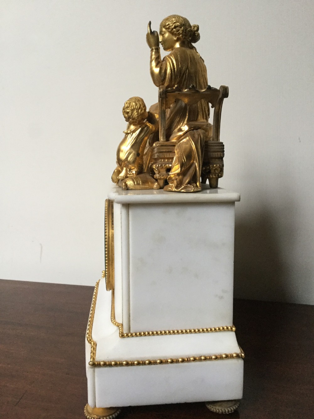 A French Neoclassical white marble mantel clock featuring a gilded bronze figural topper of a seated woman holding an open book alongside a cherub. View VI