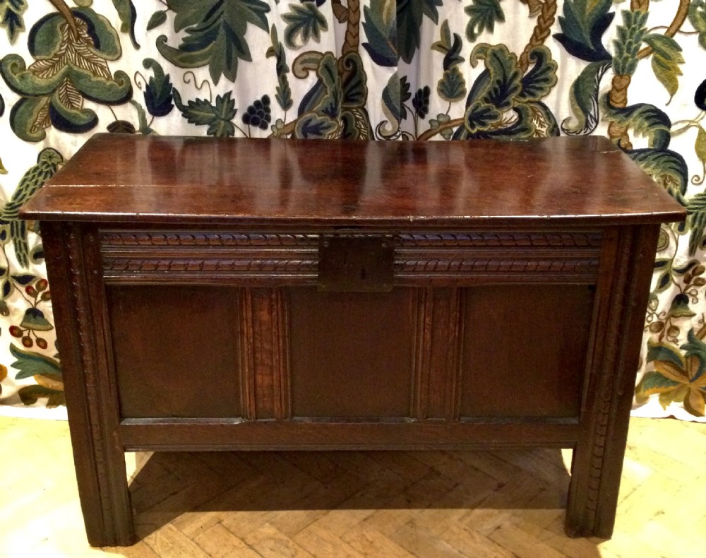 A dark carved oak coffer featuring three front panels, carved frieze detailing, an iron lock plate, and straight stile legs.