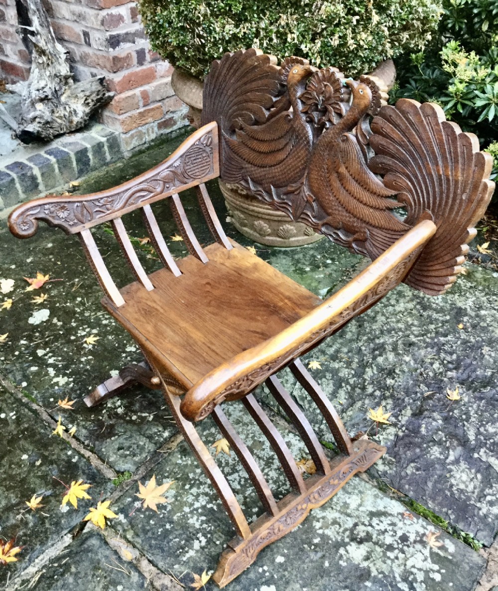 A carved brown hardwood Savonarola chair featuring an X-frame base and a backrest depicting two carved peacocks.