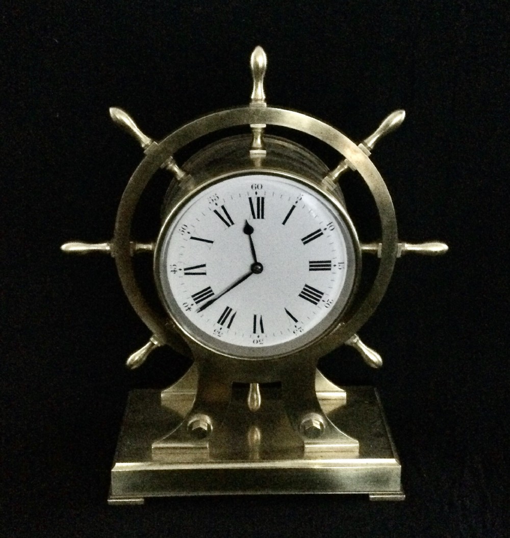 An antique brass mantel clock shaped as a ships wheel mounted on a rectangular brass base with a white Roman numeral dial.