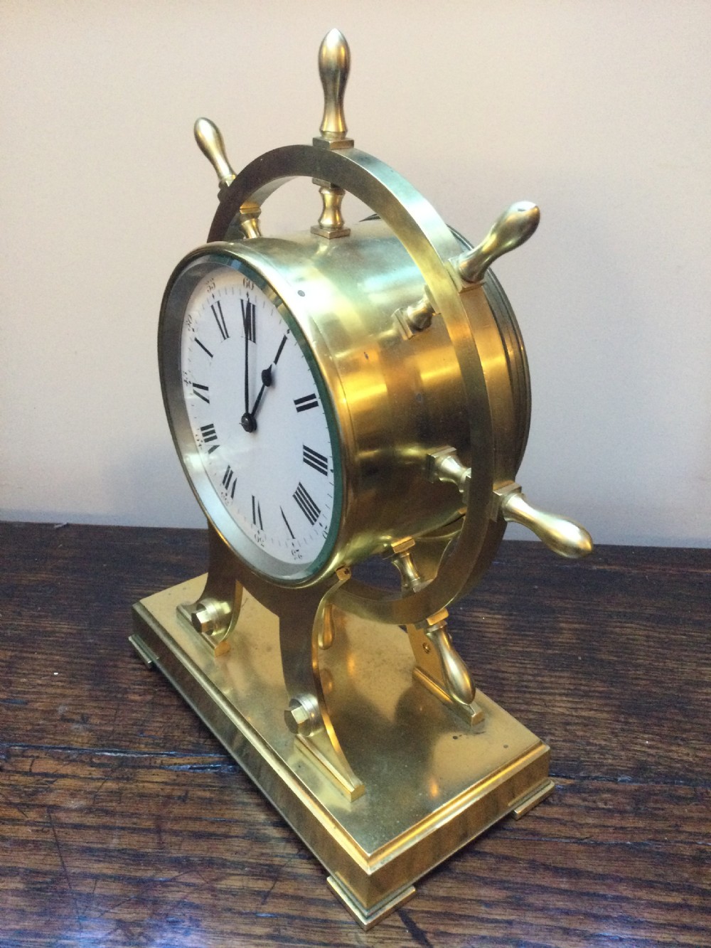 An antique brass mantel clock shaped as a ships wheel mounted on a rectangular brass base with a white Roman numeral dial. View XIV