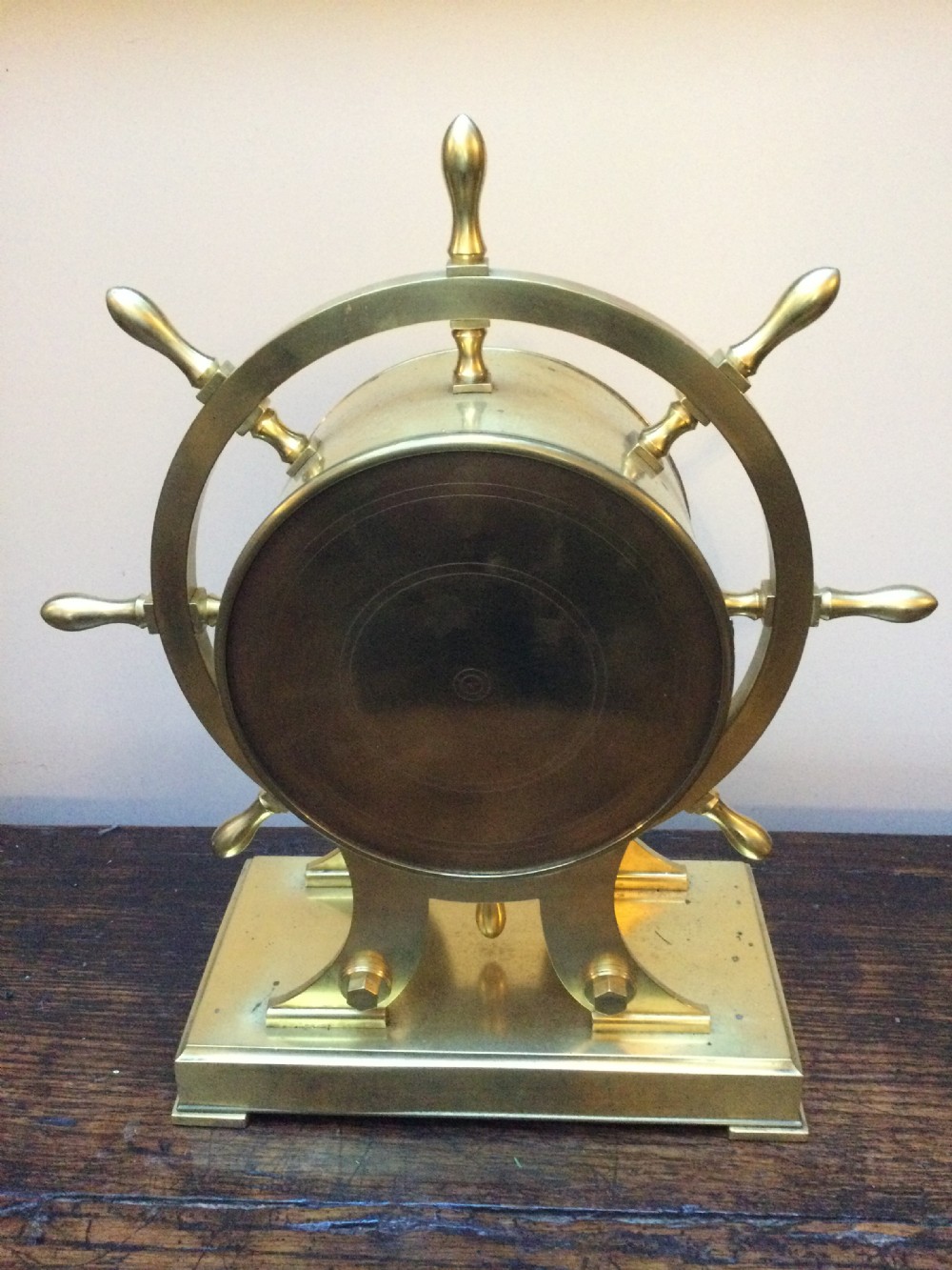 An antique brass mantel clock shaped as a ships wheel mounted on a rectangular brass base with a white Roman numeral dial. View XV