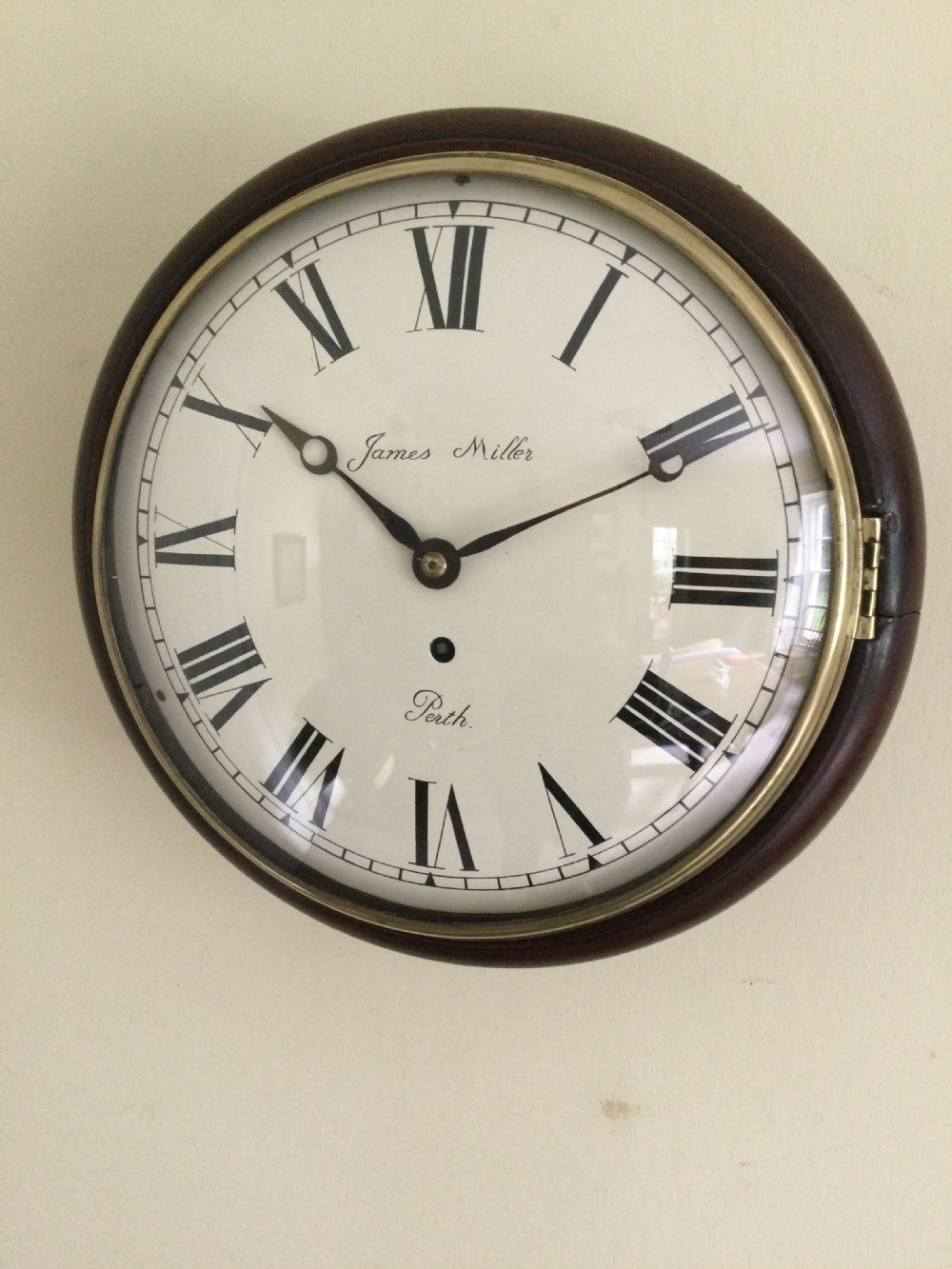 A circular antique wall clock featuring a dark mahogany case, a brass bezel, and a cream dial with black Roman numerals signed James Miller Perth. View XII