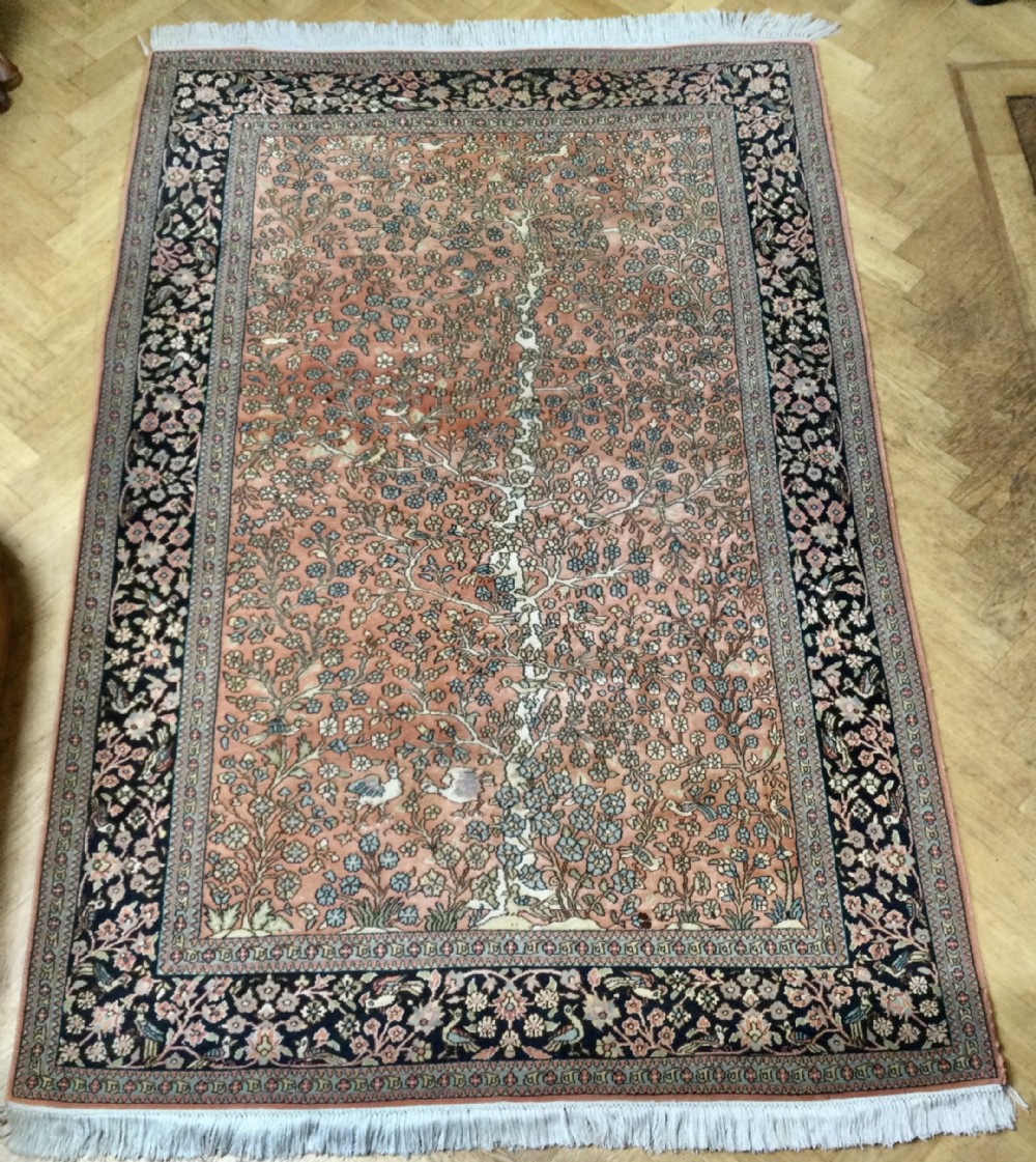 A rectangular hand-knotted Persian rug featuring a central terracotta field filled with a detailed Tree of Life and bird pattern framed by a dark navy floral border.
