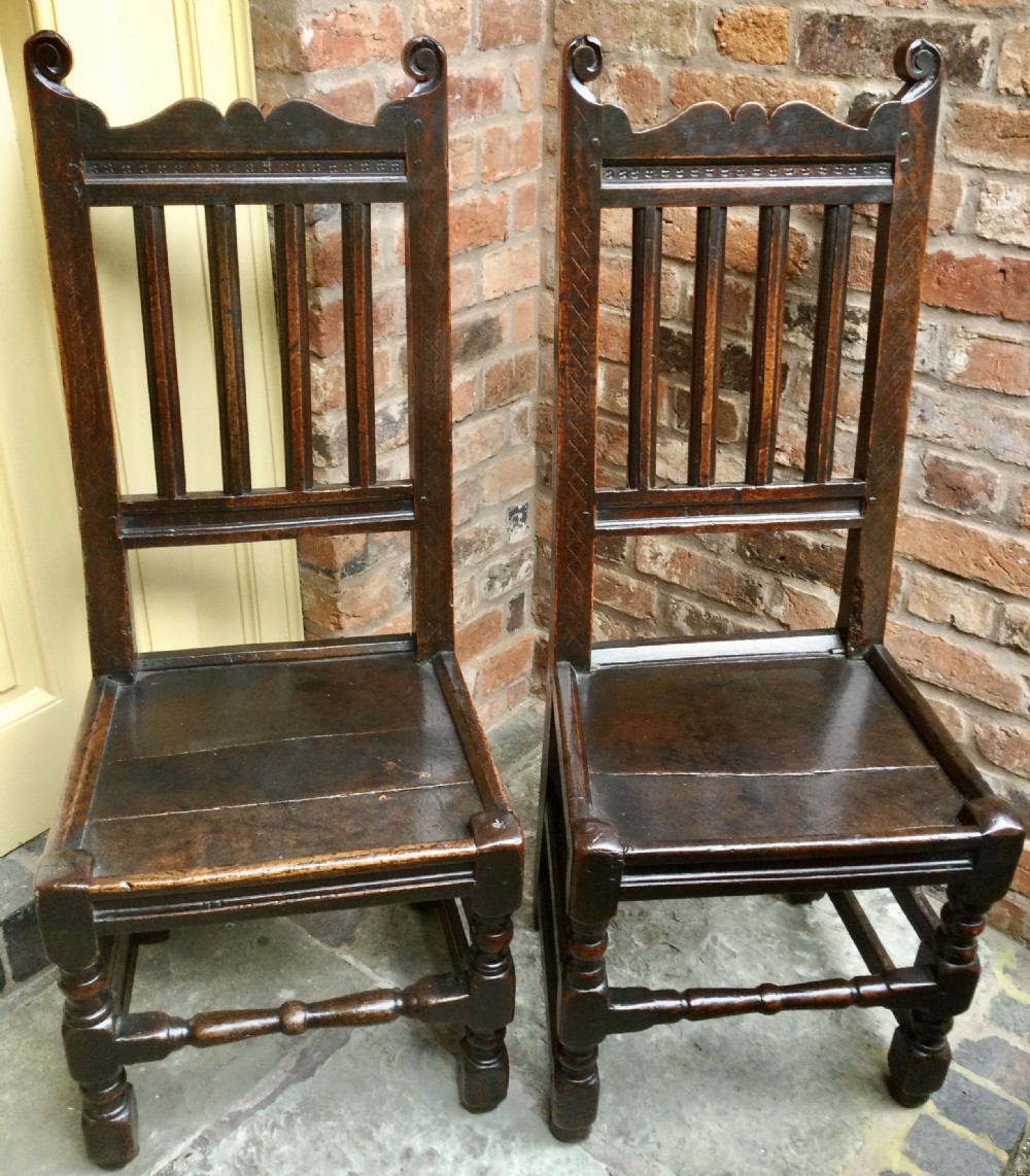 A pair of dark carved oak high-backed side chairs featuring scrolled finials, vertical back slats, solid wooden seats, and turned front legs.