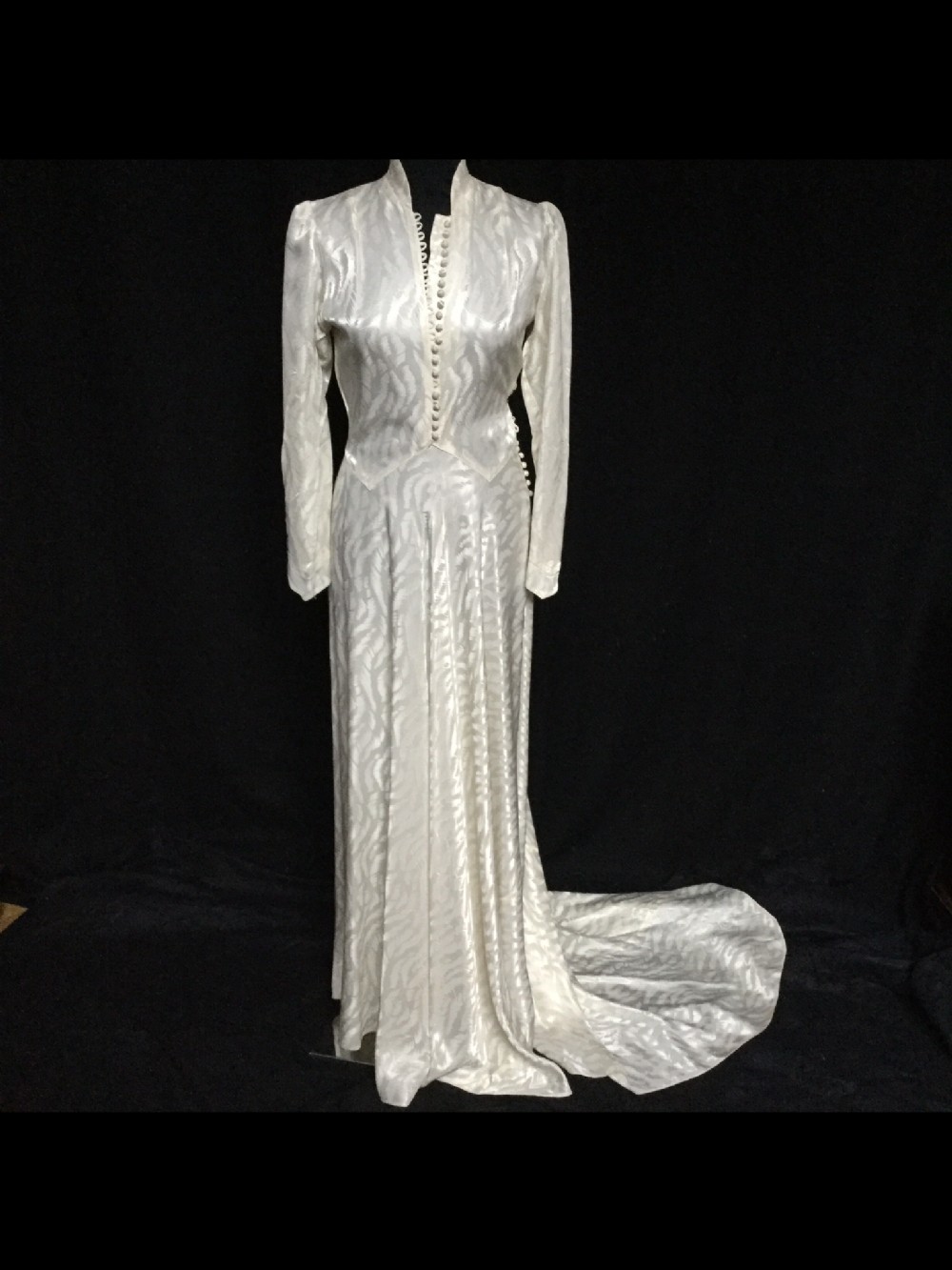 An ivory satin long-sleeved vintage wedding dress featuring a patterned jacquard weave, button-front bodice, pointed waistline, and an extended train.