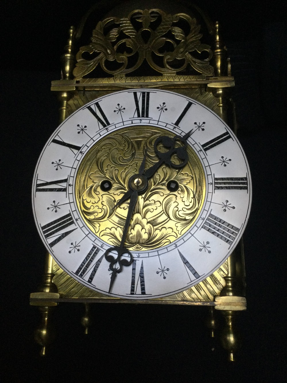 An antique brass lantern clock featuring a top bell with finial, pierced floral fretwork, and an engraved dial plate with Roman numerals. View XIV