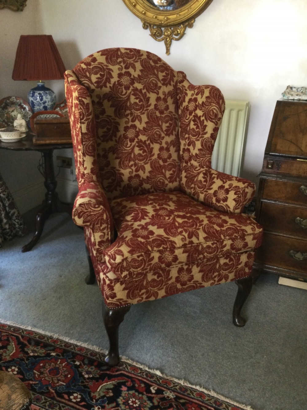 A traditional high-backed wing armchair upholstered in red and beige floral damask fabric with dark wooden cabriole legs.