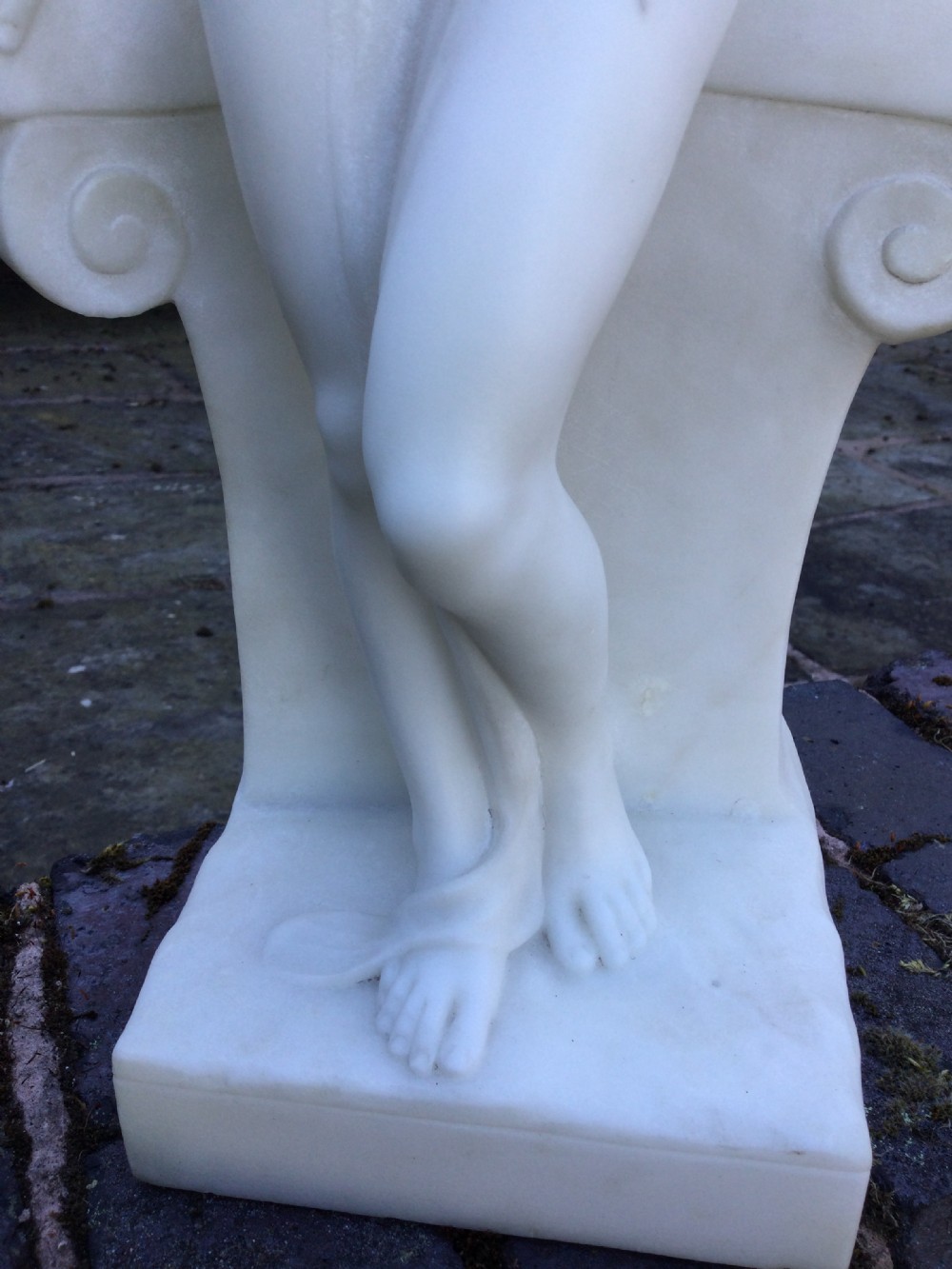 A carved off-white marble sculpture depicting a standing nude female figure leaning against a small classical ionic column base. View V