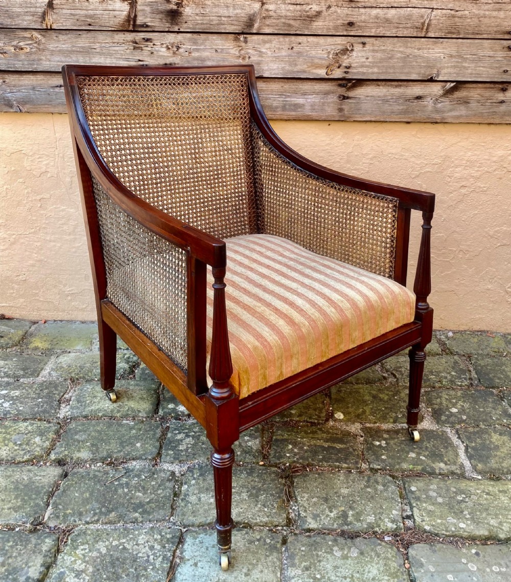 An antique mahogany armchair featuring woven cane back and side panels, turned front legs with brass castors, and a striped fabric seat cushion.