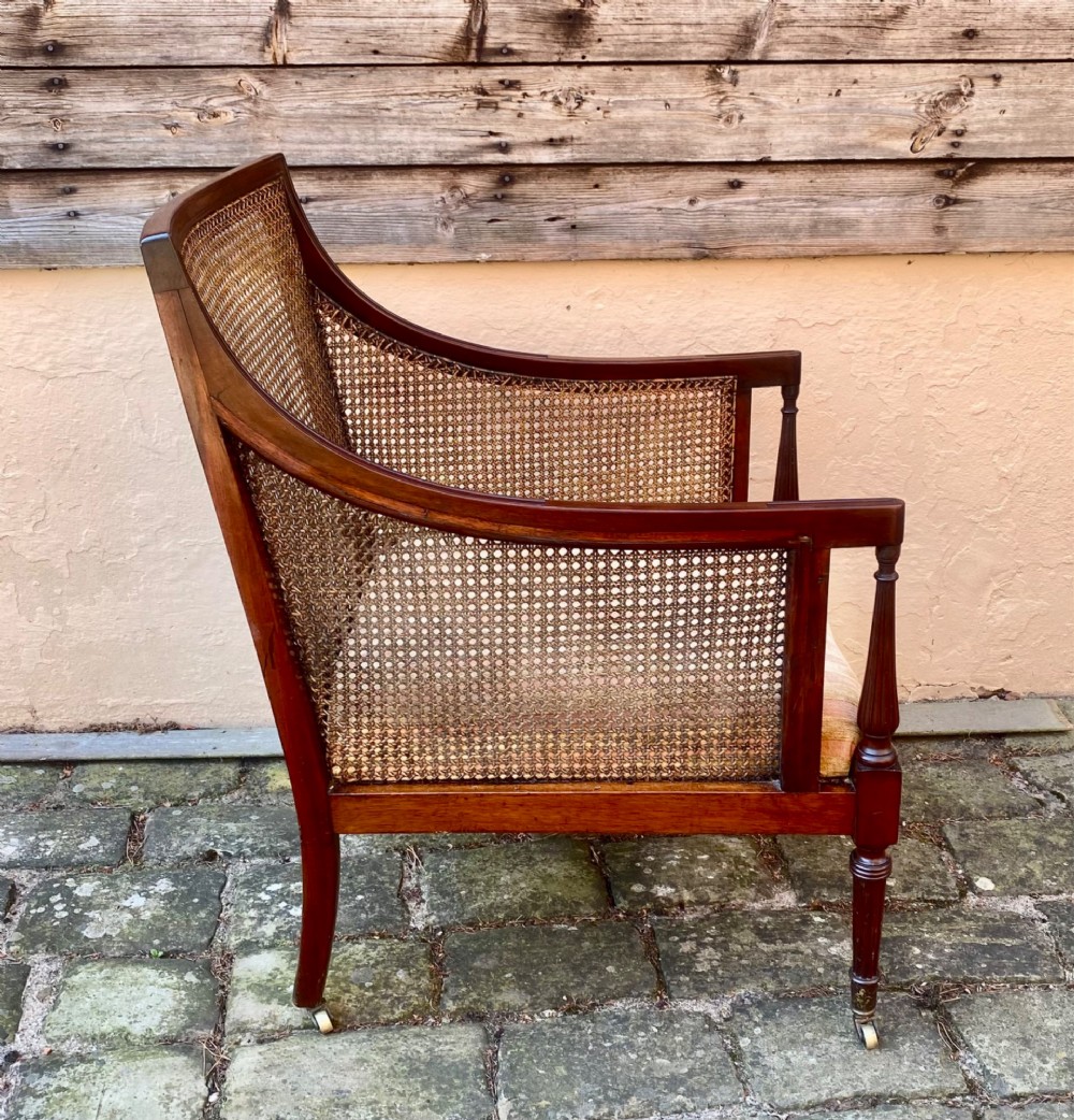 An antique mahogany armchair featuring woven cane back and side panels, turned front legs with brass castors, and a striped fabric seat cushion. View IV