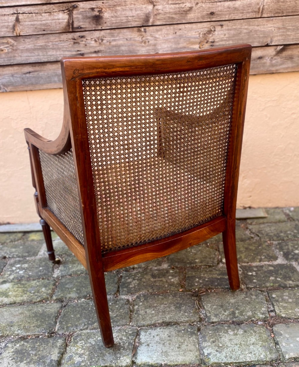 An antique mahogany armchair featuring woven cane back and side panels, turned front legs with brass castors, and a striped fabric seat cushion. View V