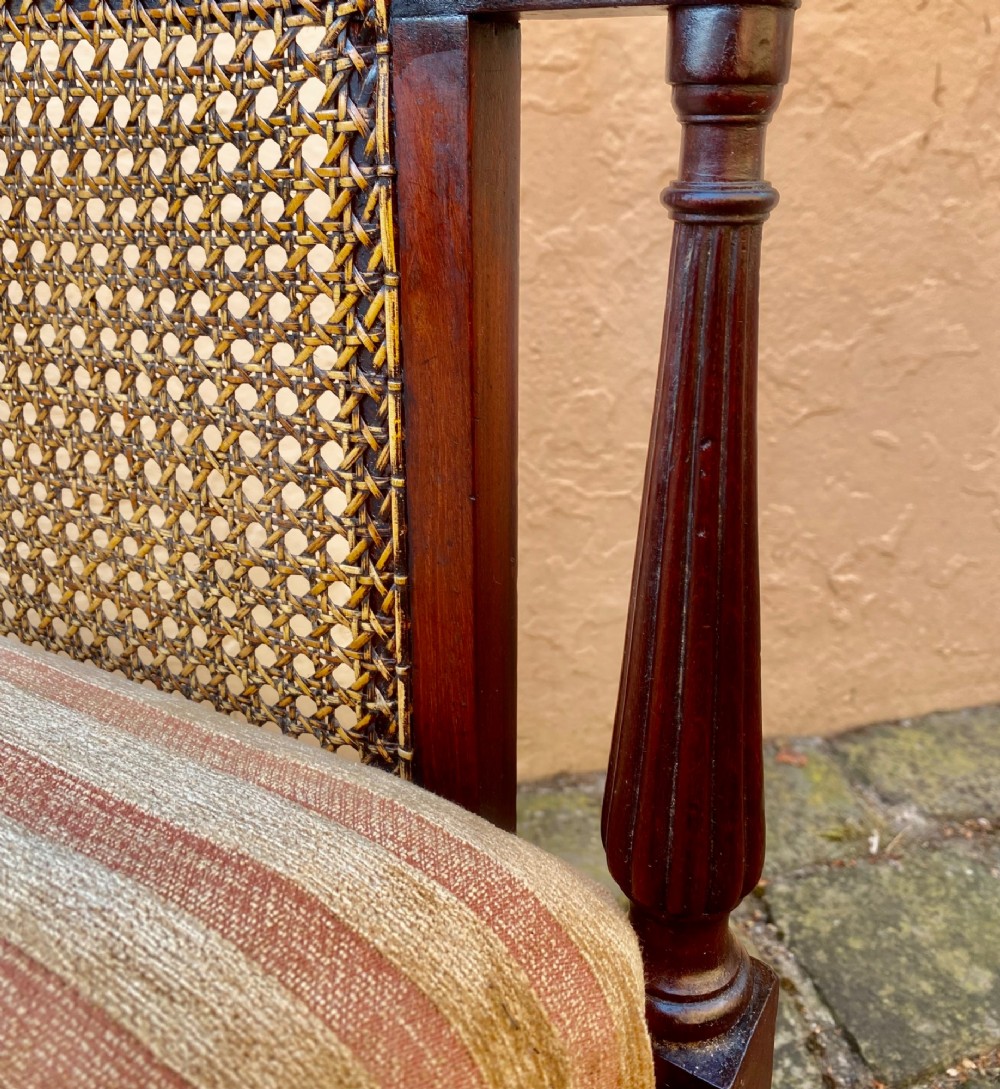 An antique mahogany armchair featuring woven cane back and side panels, turned front legs with brass castors, and a striped fabric seat cushion. View VIII