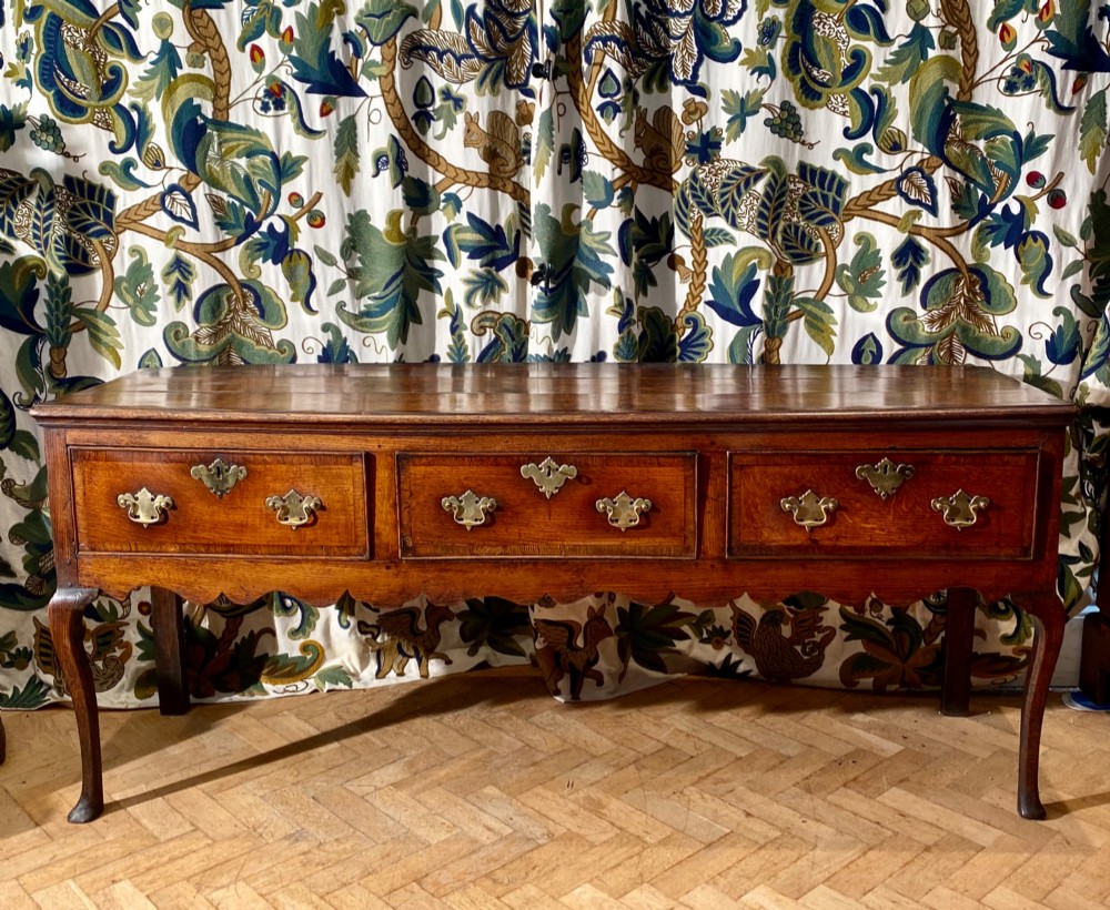 A Georgian oak dresser base featuring three drawers with brass handles, a scalloped apron, and front cabriole legs.