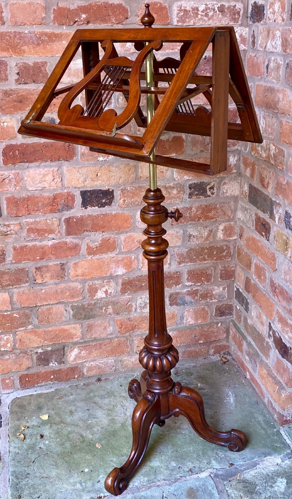An antique mahogany duet music stand featuring double lyre-shaped rests, an adjustable brass stem, and a turned pedestal base on tripod feet.