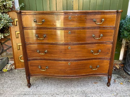 fig 1. A George III Sheraton period mahogany chest of drawers featuring a curved serpentine front, four graduated drawers, brass swan-neck handles, and reeded corner columns.