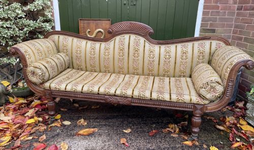 fig 1. An early nineteenth-century Regency mahogany sofa featuring double scroll-ended arms, a carved crest rail, striped gold velvet upholstery, matching bolster cushions, and turned legs on brass castors.