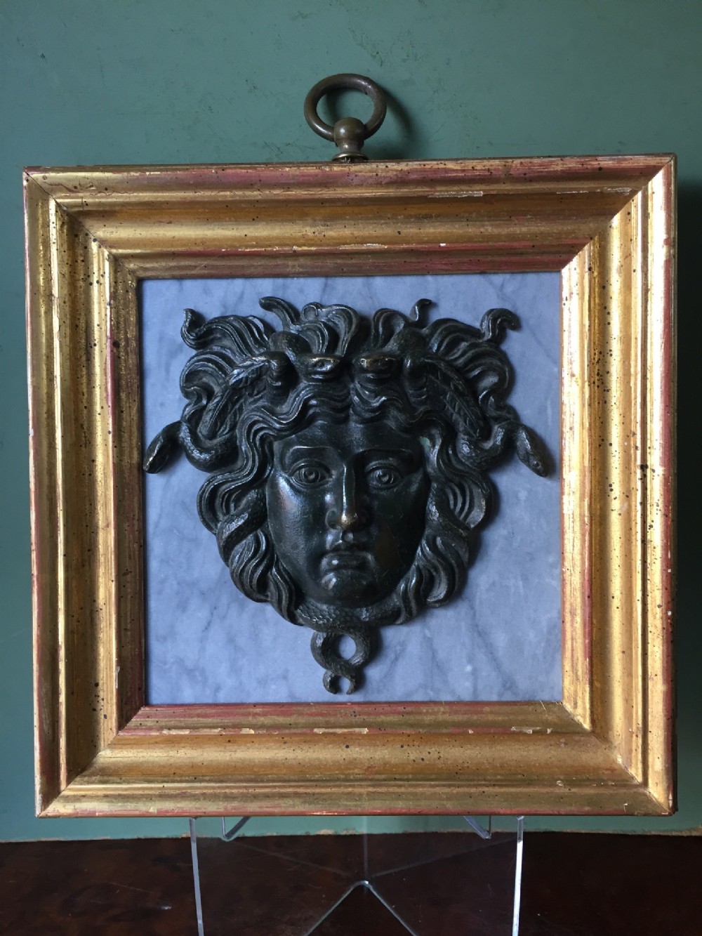 C19th 'Grand Tour' souvenir bronze mask of Medusa , mounted upon a pale grey marble panel and set in a gilt-wood frame.