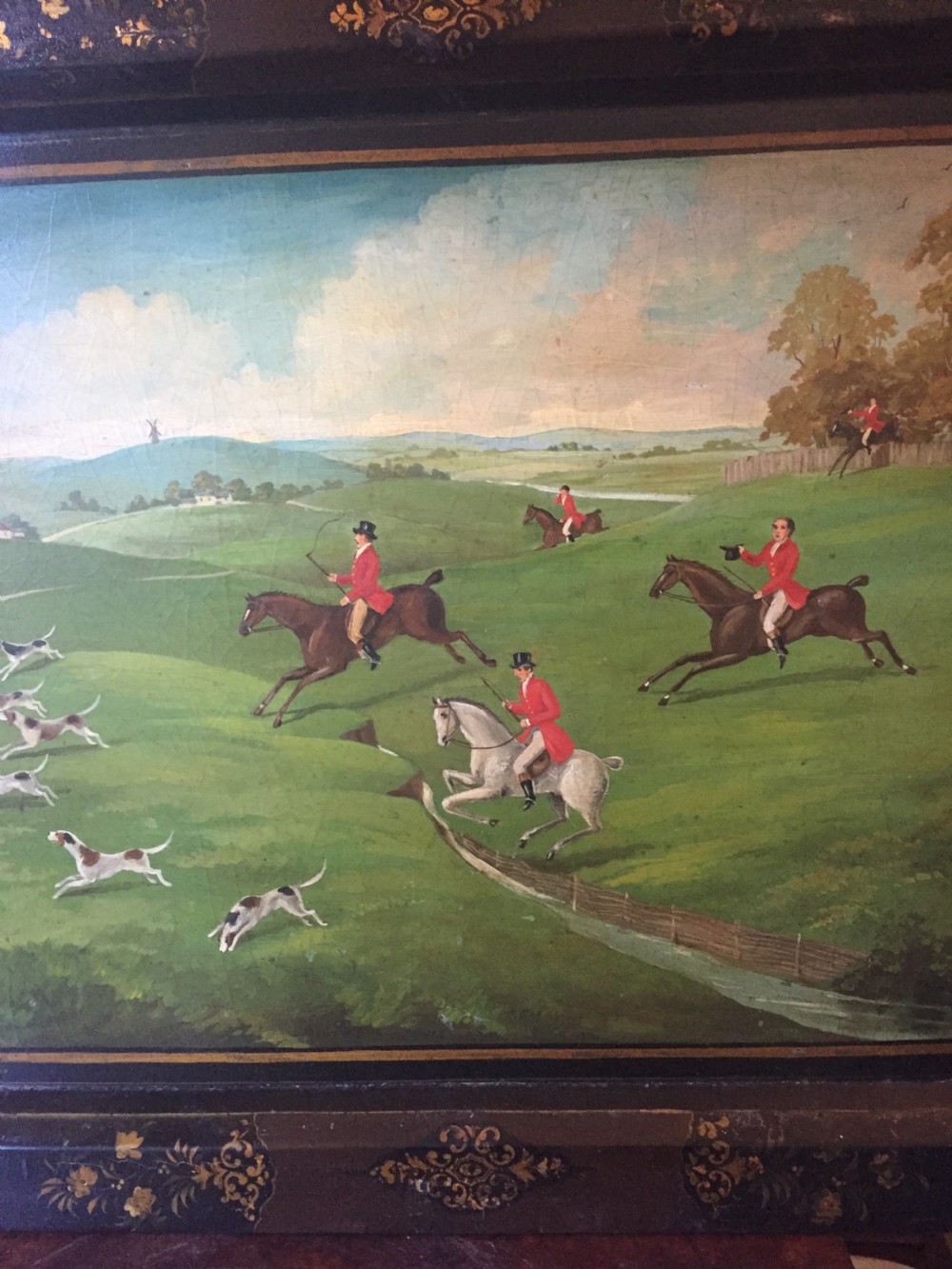 An early 19th-century rectangular papier-mache tray featuring a gilded scrollwork border and a central hand-painted scene of horsemen and hounds engaged in a fox hunt across a countryside landscape. View V