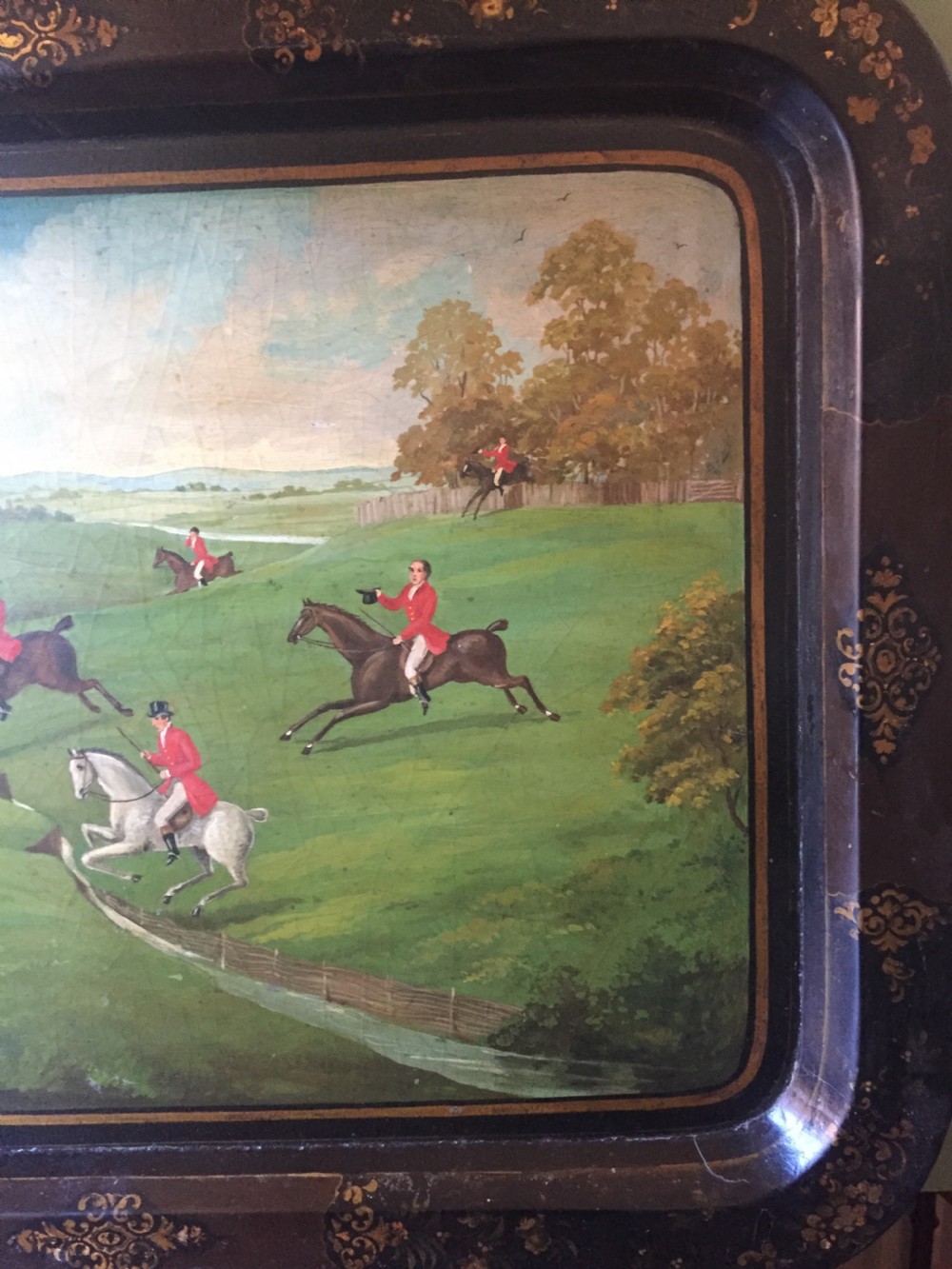 An early 19th-century rectangular papier-mache tray featuring a gilded scrollwork border and a central hand-painted scene of horsemen and hounds engaged in a fox hunt across a countryside landscape. View VI
