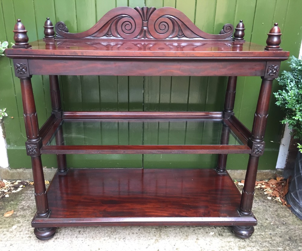 A George IV mahogany three-tier buffet featuring a carved scroll upstand, turned finials, column supports, and a glass-inset middle shelf.