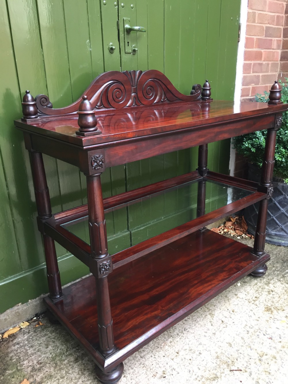 A George IV mahogany three-tier buffet featuring a carved scroll upstand, turned finials, column supports, and a glass-inset middle shelf. View III