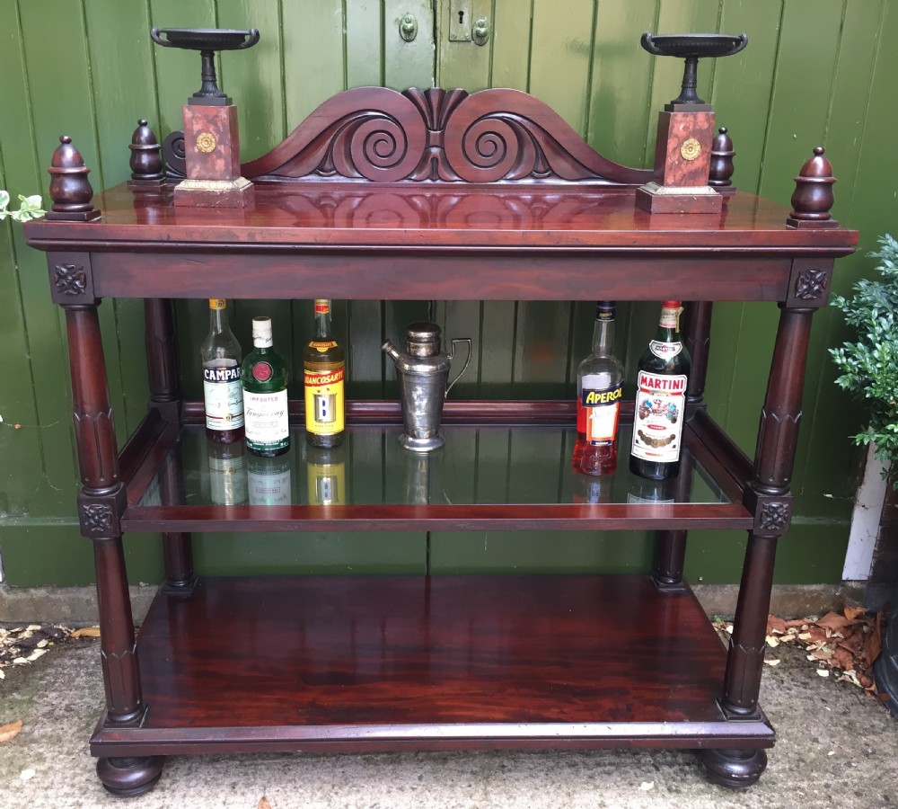 A George IV mahogany three-tier buffet featuring a carved scroll upstand, turned finials, column supports, and a glass-inset middle shelf. View XVI