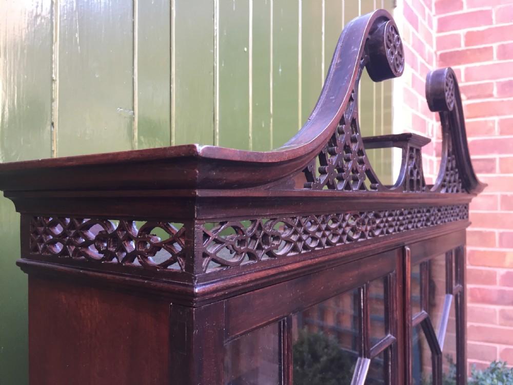 A late 19th-century Chippendale Revival mahogany glazed cabinet on stand featuring a swan-neck pediment, fretwork frieze, astragal glass doors, and turned legs with a lower shelf. View X