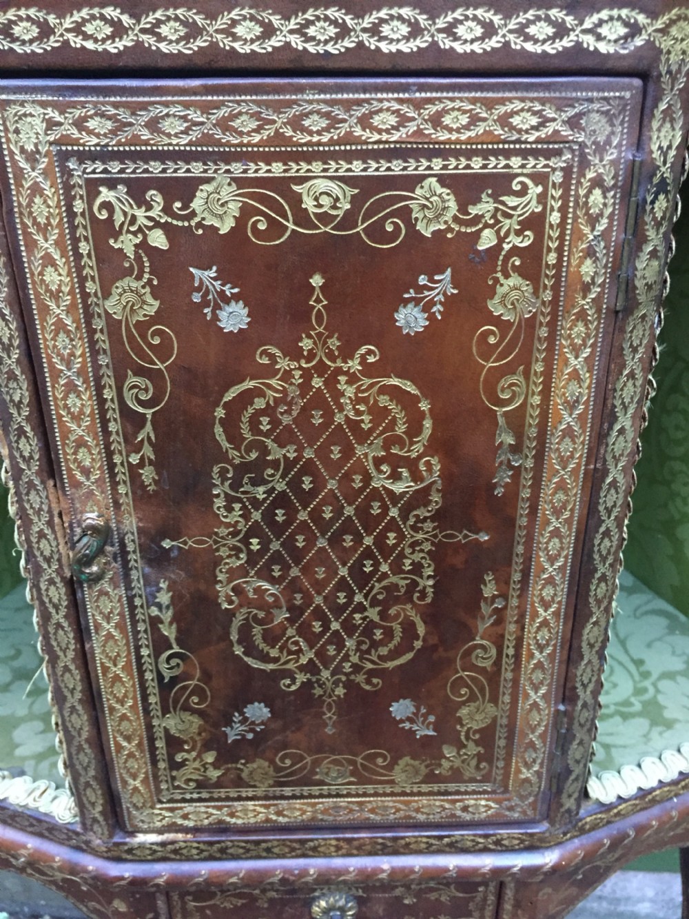 An early twentieth-century French accent table encased in chestnut brown leather with gold leaf tooling, featuring a central cupboard door, open side compartments lined in green damask silk, a small drawer, and cabriole legs. View X