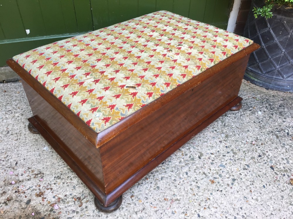 A mid-19th-century mahogany storage ottoman featuring a geometric gros-point needlework upholstered top resting on low turned bun feet.