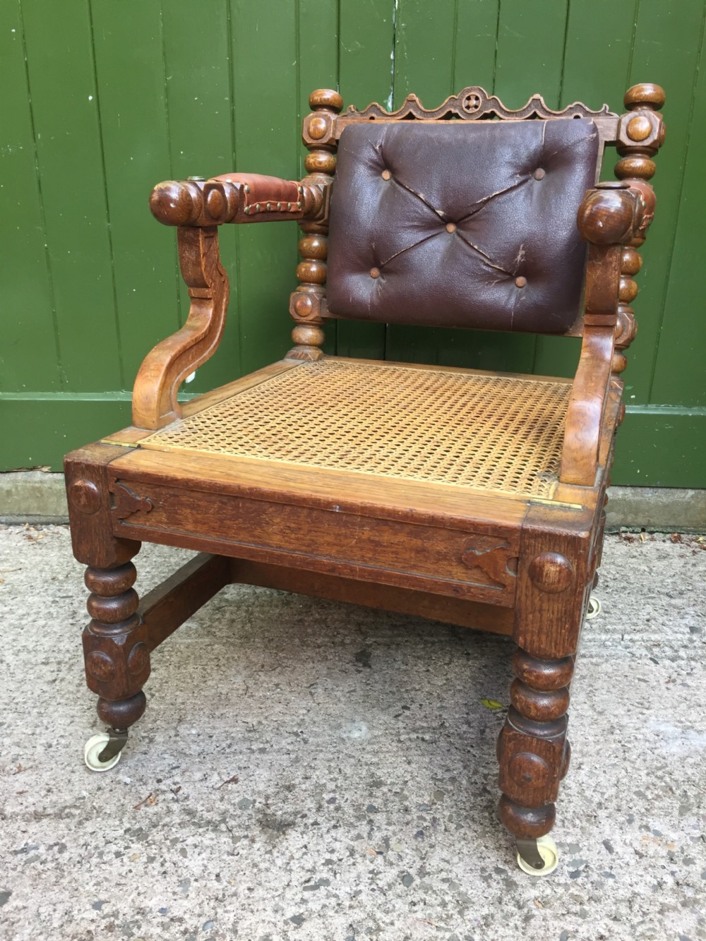 An early nineteenth-century carved oak metamorphic library armchair featuring a button-tufted leather backrest, caned seat, bobbin-turned details, and brass casters. View XVII