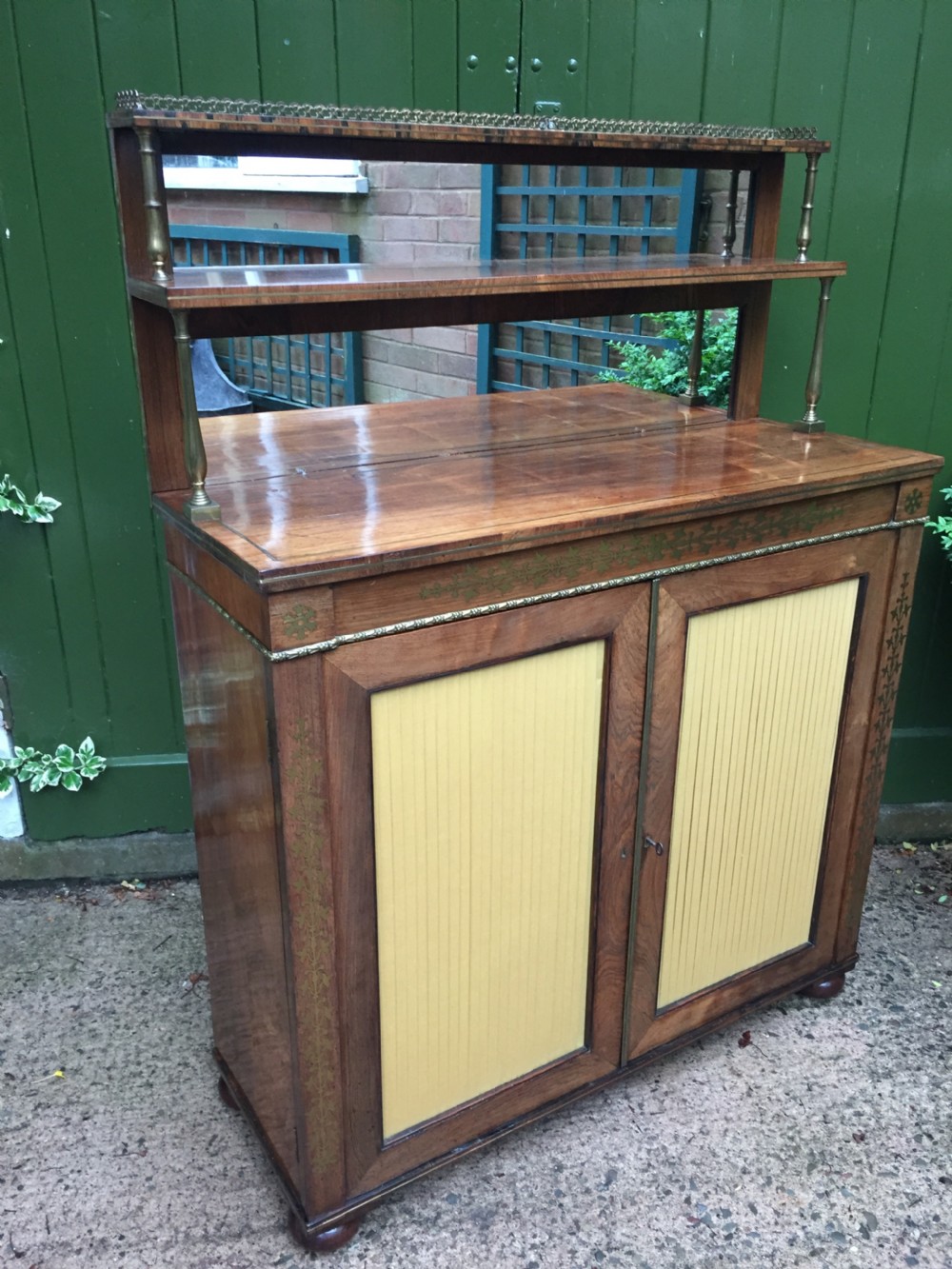An early nineteenth-century Regency rosewood chiffonier featuring brass floral inlay, an upper shelf supported by brass columns, and twin cabinet doors with yellow pleated silk panels. View II