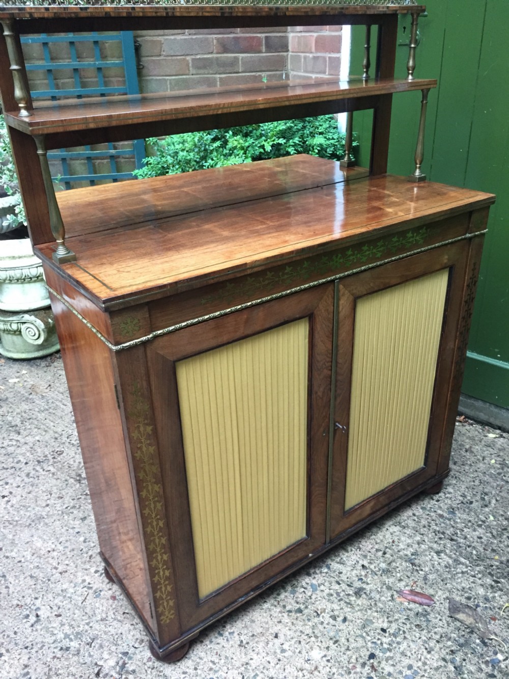 An early nineteenth-century Regency rosewood chiffonier featuring brass floral inlay, an upper shelf supported by brass columns, and twin cabinet doors with yellow pleated silk panels. View III