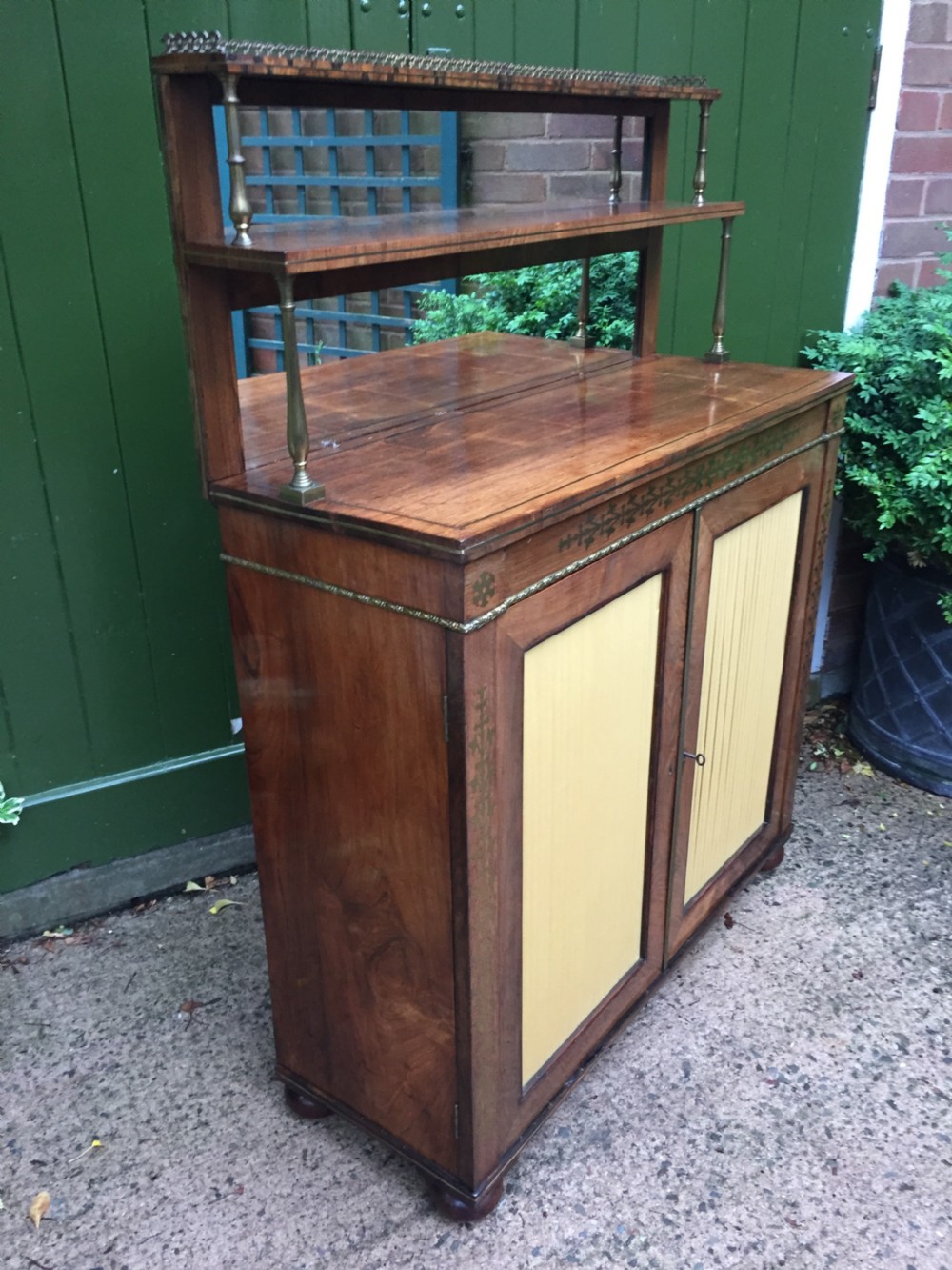 An early nineteenth-century Regency rosewood chiffonier featuring brass floral inlay, an upper shelf supported by brass columns, and twin cabinet doors with yellow pleated silk panels. View V