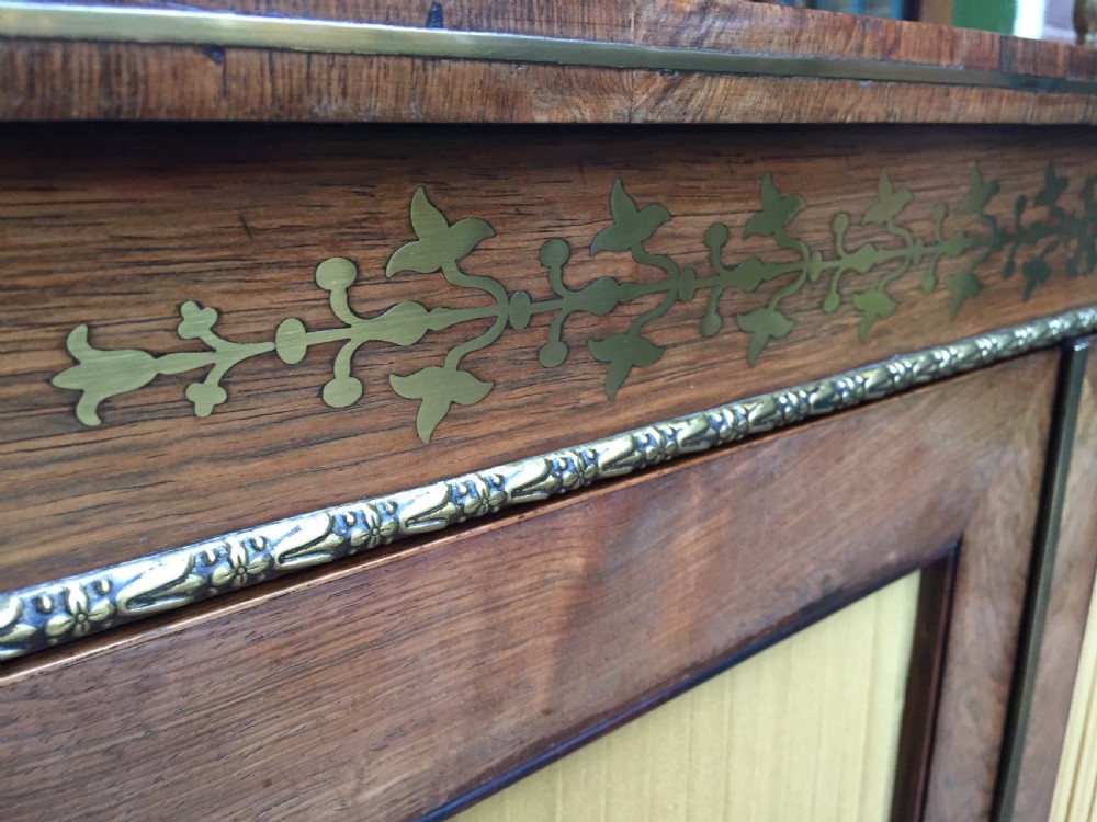 An early nineteenth-century Regency rosewood chiffonier featuring brass floral inlay, an upper shelf supported by brass columns, and twin cabinet doors with yellow pleated silk panels. View X