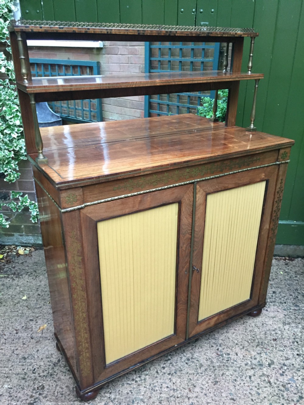 An early nineteenth-century Regency rosewood chiffonier featuring brass floral inlay, an upper shelf supported by brass columns, and twin cabinet doors with yellow pleated silk panels. View XIV