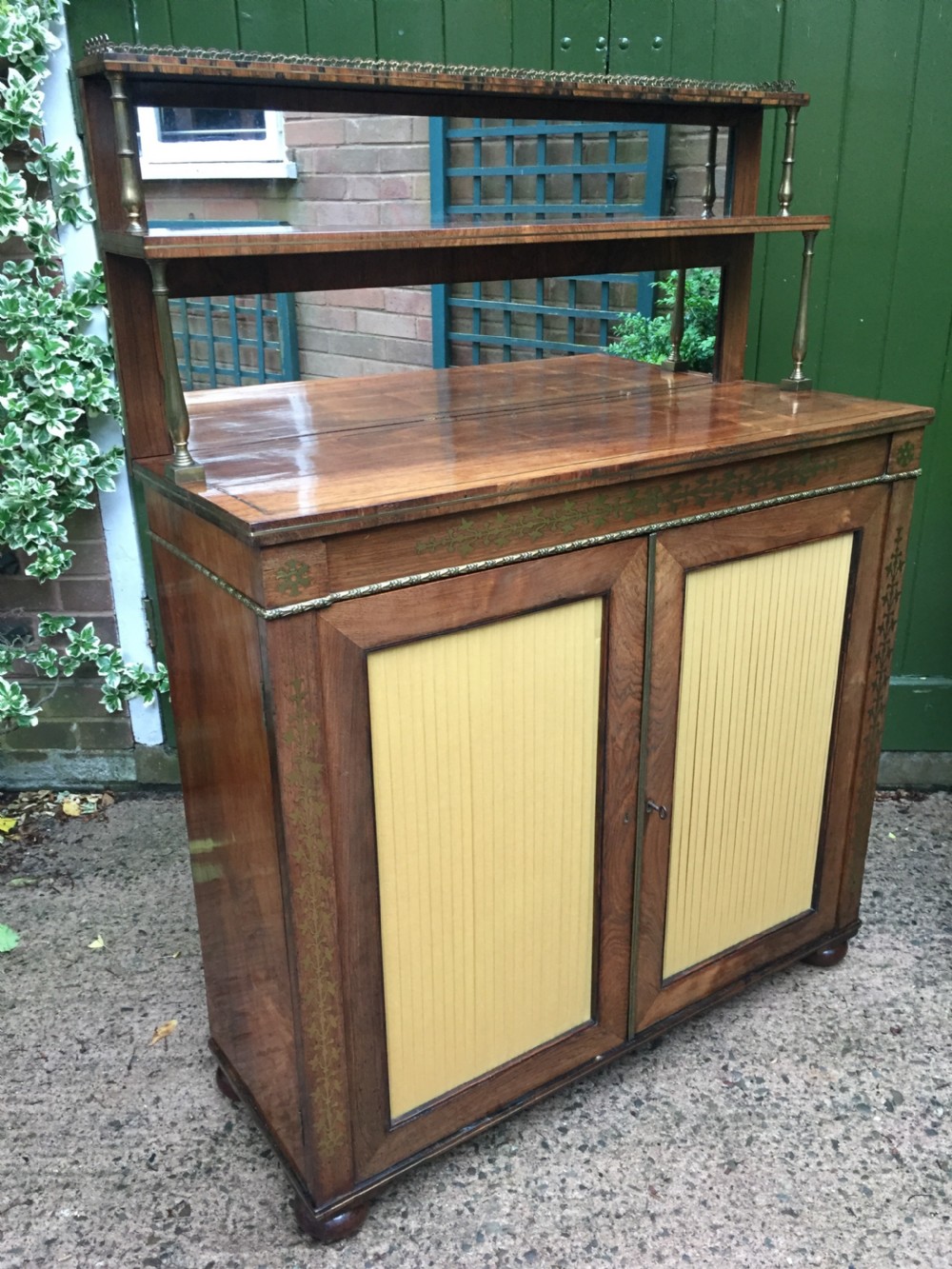 An early nineteenth-century Regency rosewood chiffonier featuring brass floral inlay, an upper shelf supported by brass columns, and twin cabinet doors with yellow pleated silk panels. View XVI