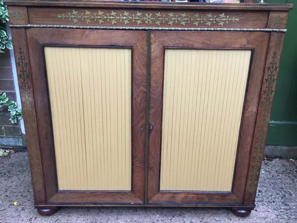 An early nineteenth-century Regency rosewood chiffonier featuring brass floral inlay, an upper shelf supported by brass columns, and twin cabinet doors with yellow pleated silk panels. View XIX