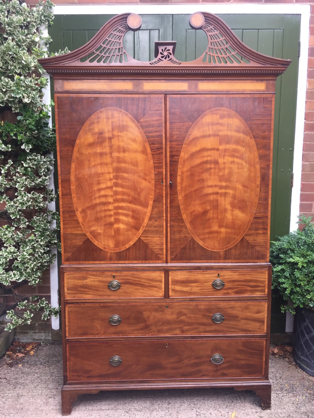 A late 18th-century George III mahogany and satinwood linen press with a swan-neck pediment, oval inlaid cupboard doors, and four lower drawers.