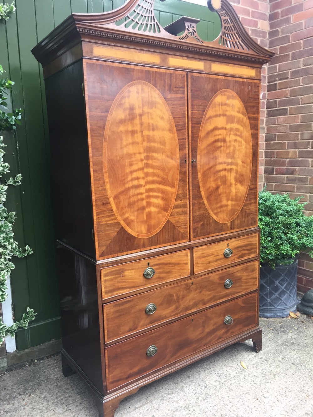 A late 18th-century George III mahogany and satinwood linen press with a swan-neck pediment, oval inlaid cupboard doors, and four lower drawers. View VI